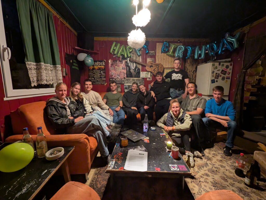 Ein Bild eines Jugendraums mit roten Wänden, einem Tisch und einer Sofaecke auf dem 11 junge Menschen sitzen und in die Kamera schauen.