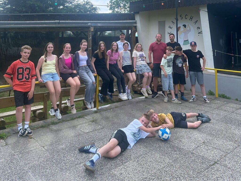 Eine Gruppe von jungen Menschen, die auf einer Stange sitzen. Im Vordergrund liegen zwei junge Menschen auf dem Boden. Alle schauen in die Kamera.