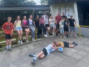 Eine Gruppe von jungen Menschen, die auf einer Stange sitzen. Im Vordergrund liegen zwei junge Menschen auf dem Boden. Alle schauen in die Kamera.