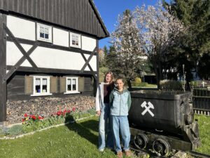 Zwei junge Projektemacherinnen vor einer Bergwerklore, die auf einer Wiese im Freien steht. An der Seite ist ein Ausschnitt eines Fachwerkhauses zu sehen.