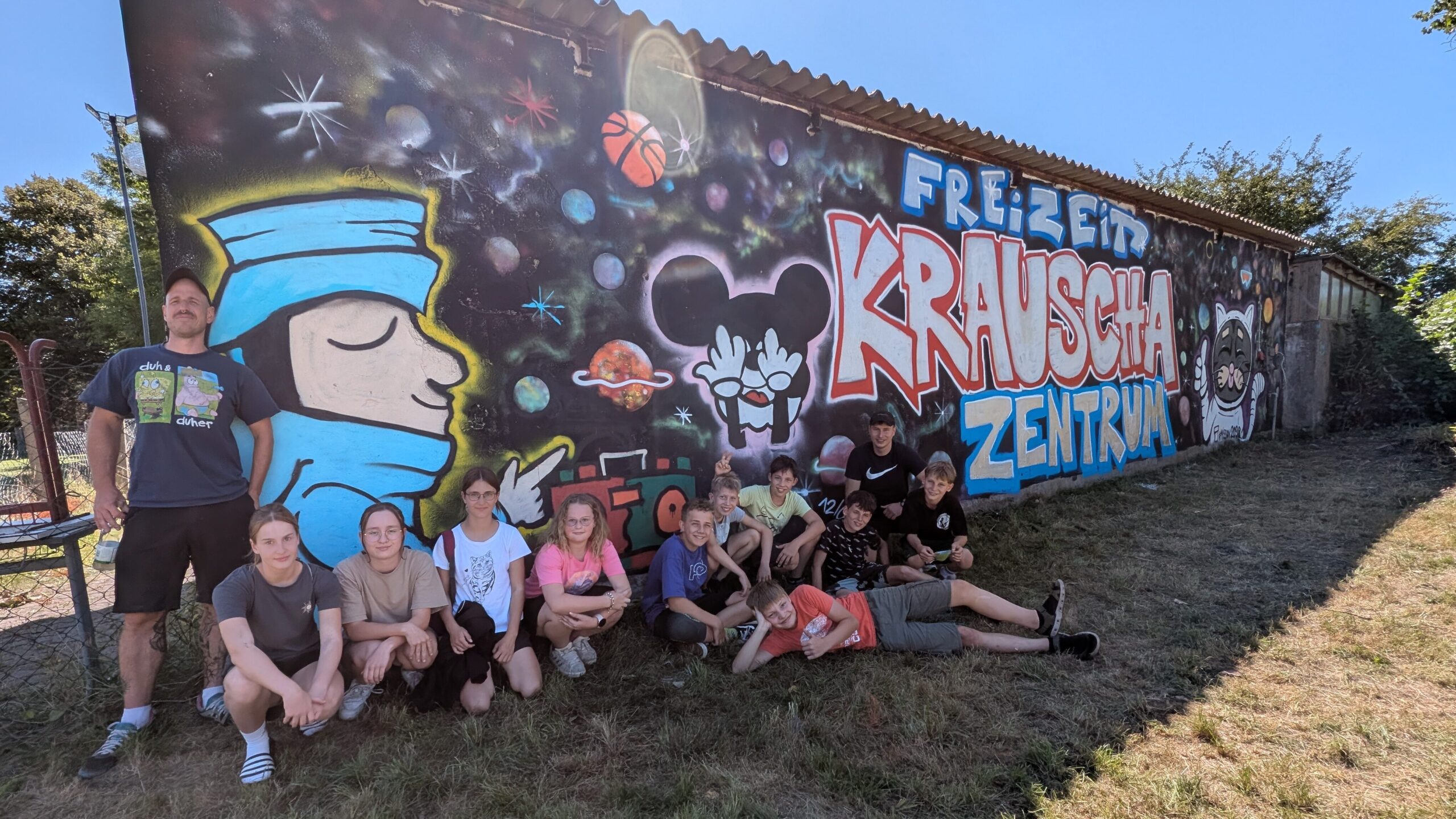 Eine Gruppe von 10 Kindern und 2 Erwachsenen, die vor einer angesprühten Fassade stehen und für ein Gruppenbild posieren. Auf der Wand ist der Schriftzug Freizeitzentrum Krauscha zu lesen. Die Wand hat einen schwarzen Hintergrund und mehrere Graffitimotive mit Bezug zum Weltraum.